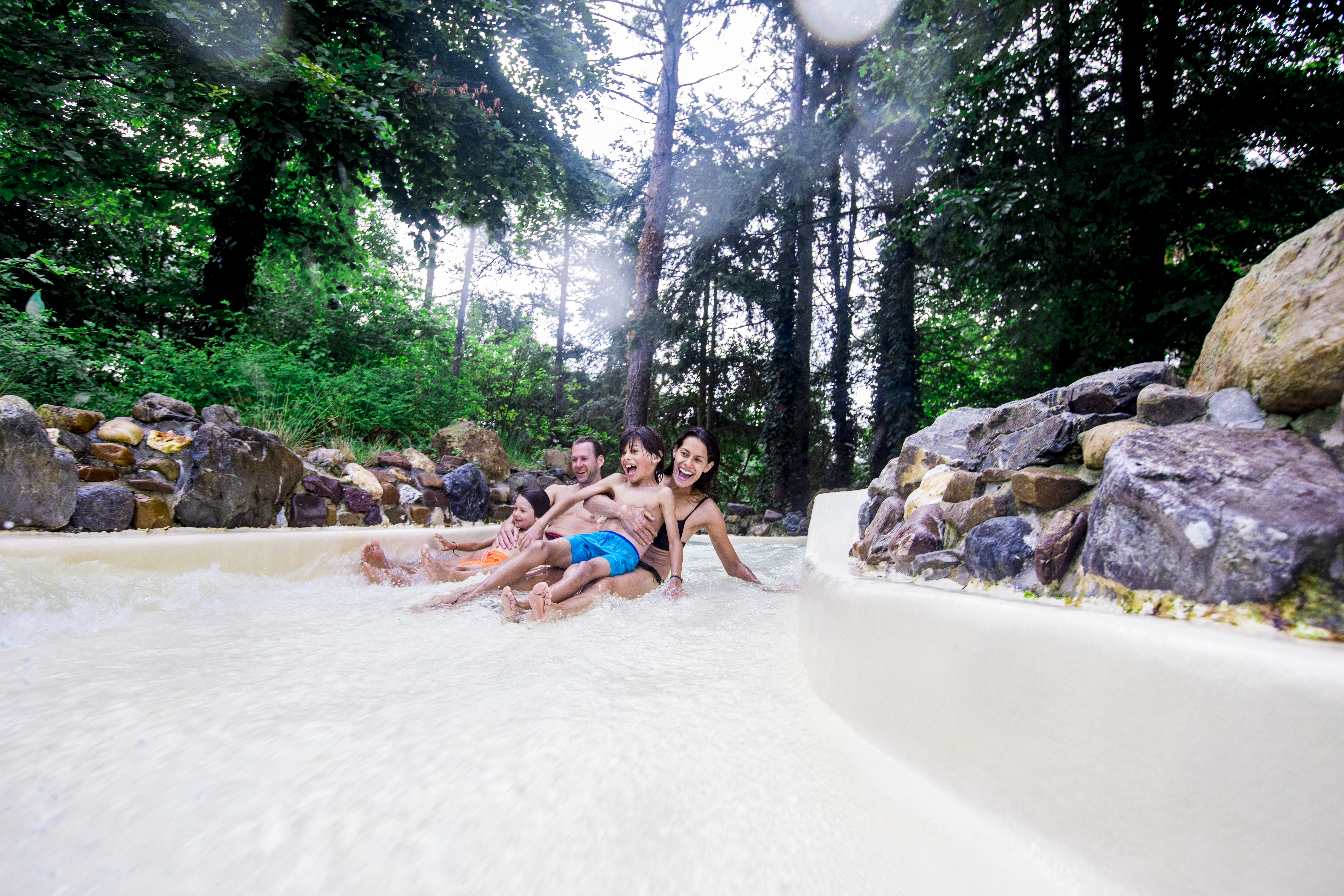 Een gezin glijdt samen door de wildwaterbaan van de Aqua Mundo bij Center Parcs Erperheide, omgeven door groene bomen en rotsen.