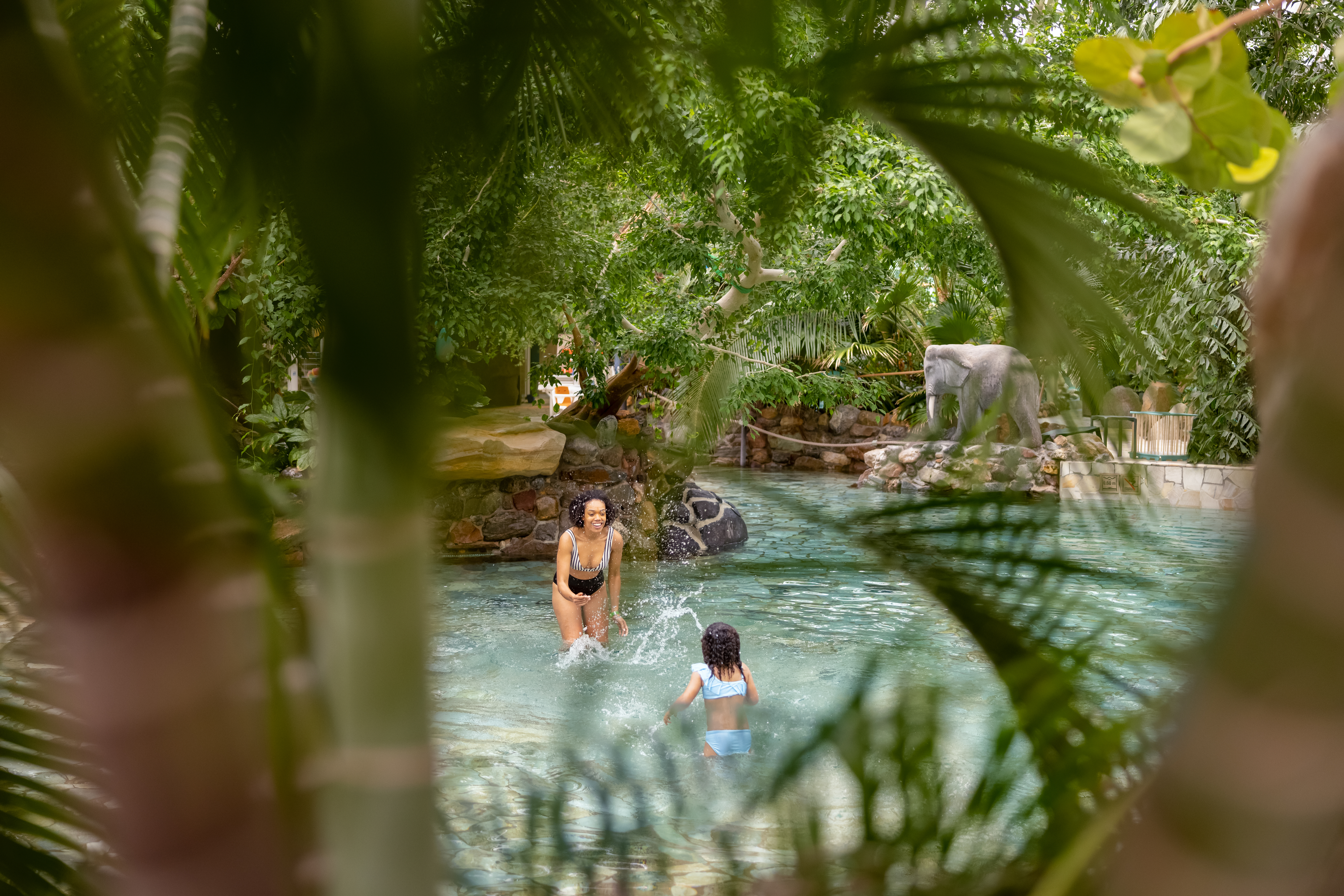 Een moeder en dochter spelen en spetteren in het water van het tropische zwembad in de Aqua Mundo van Center Parcs Erperheide, omgeven door weelderige planten.