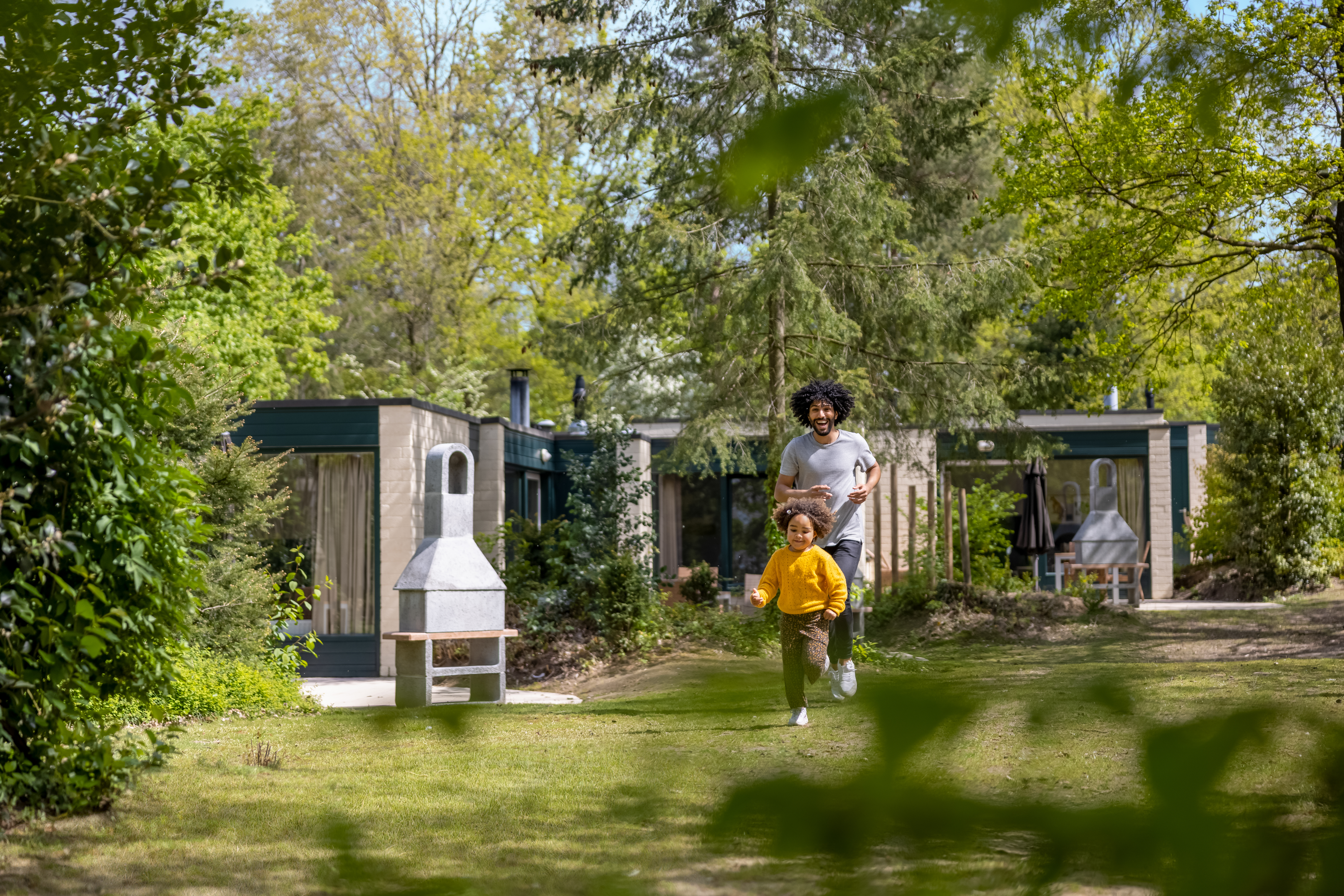 Een kind rent vrolijk door de ruime, groene tuin van een cottage bij Center Parcs Erperheide, met een barbecue op het terras.