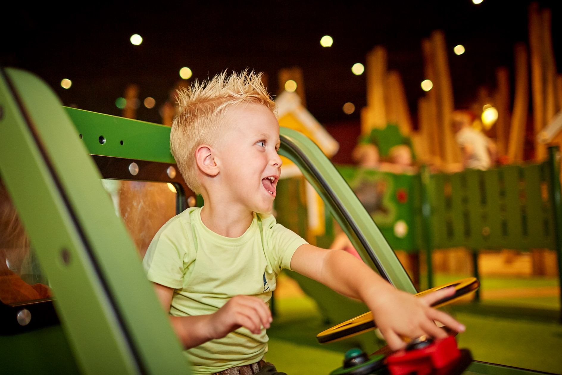 Een lachende jongen met blond haar zit in een groen speeltoestel en drukt op een knop in de indoor speeltuin La Selva op De Leistert.