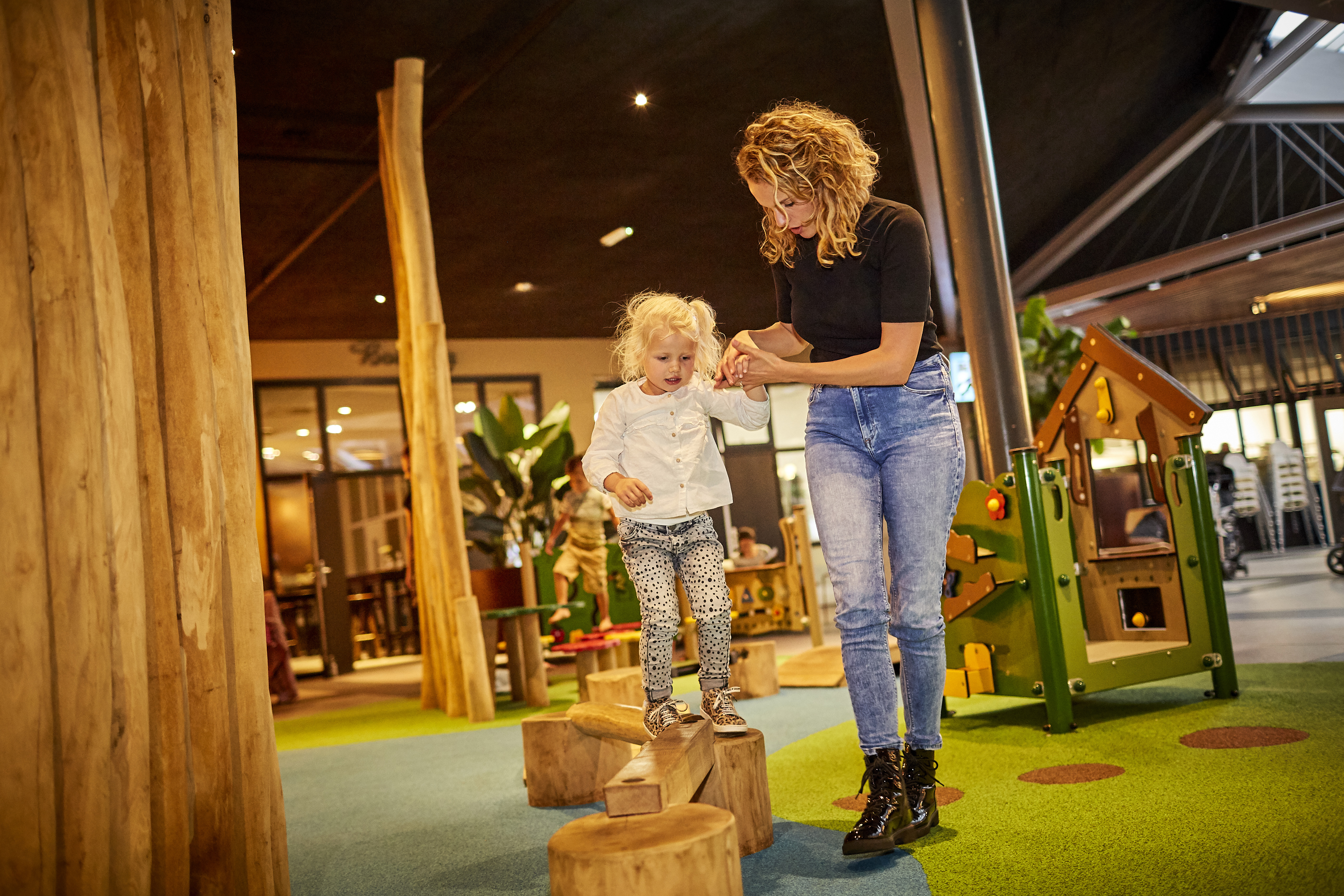 Een moeder helpt haar jonge dochter met balanceren op houten paaltjes in het peutergedeelte van de indoor speeltuin op Recreatiepark De Leistert.