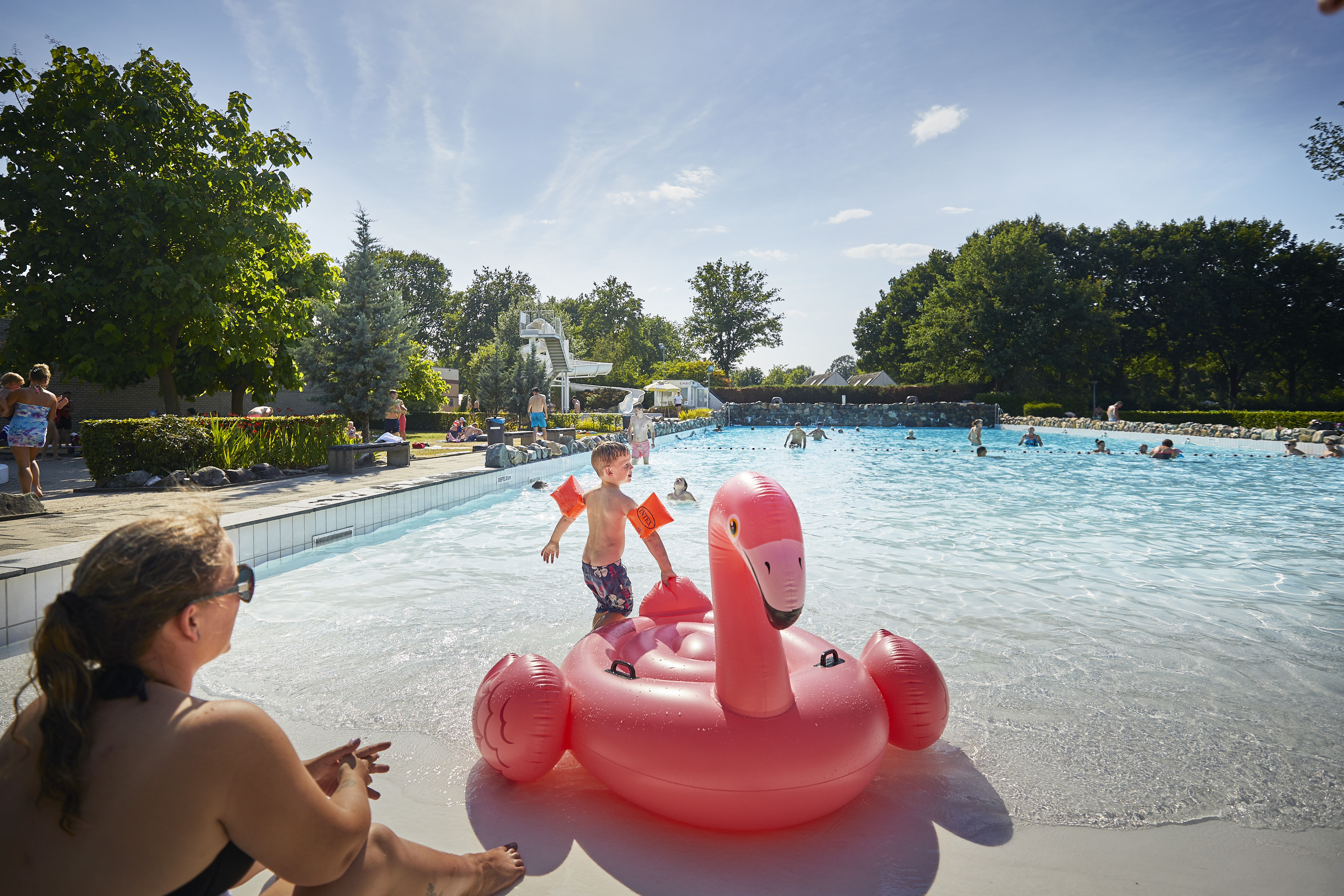 Een kind speelt met een opblaasbare roze flamingo in het buitengolfslagbad van Recreatiepark De Leistert op een zonnige dag.