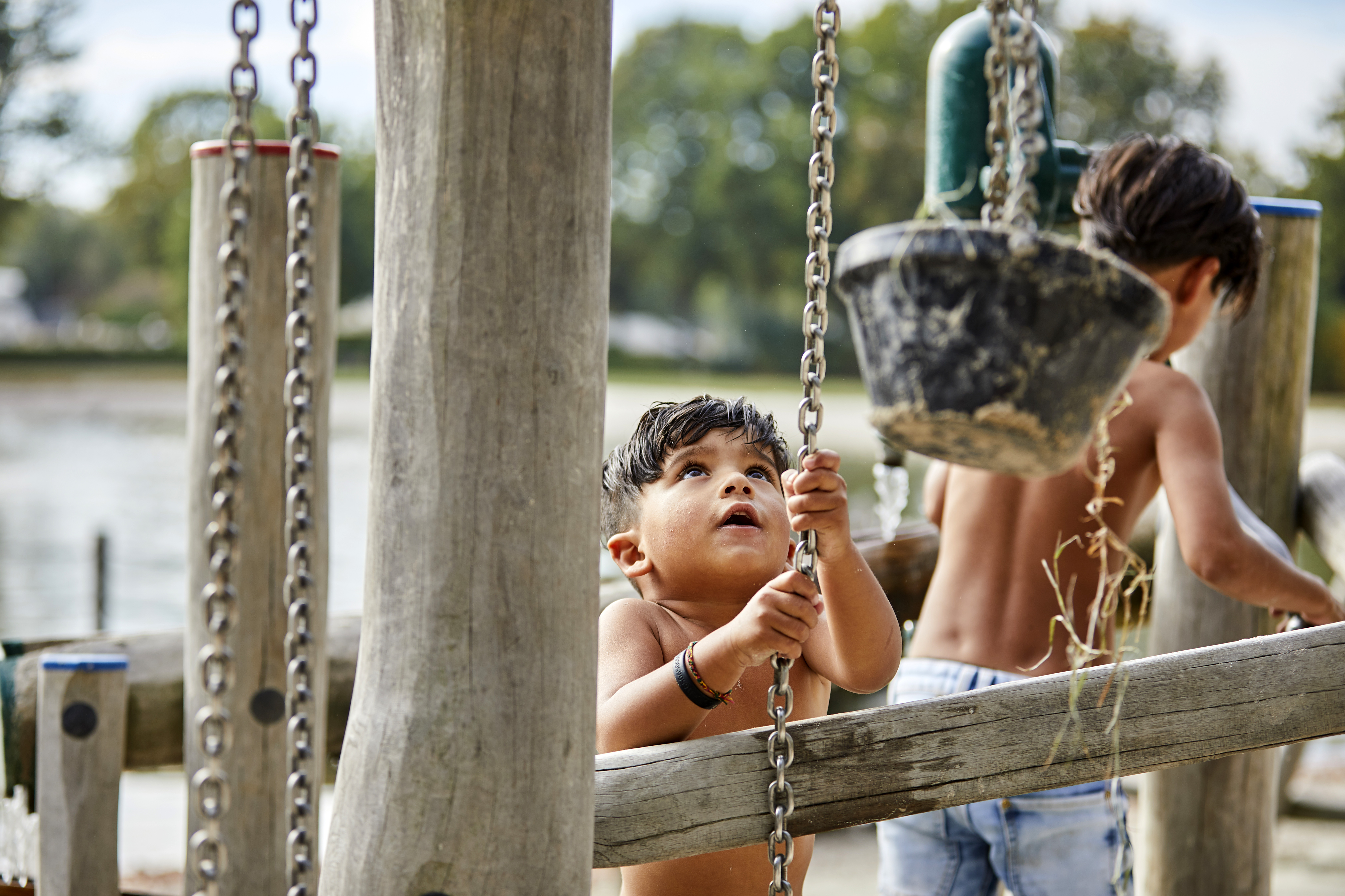 Twee jonge kinderen spelen met water en zand bij een houten speeltoestel met emmertjes en kettingen in de waterspeeltuin aan het strand van De Leistert.