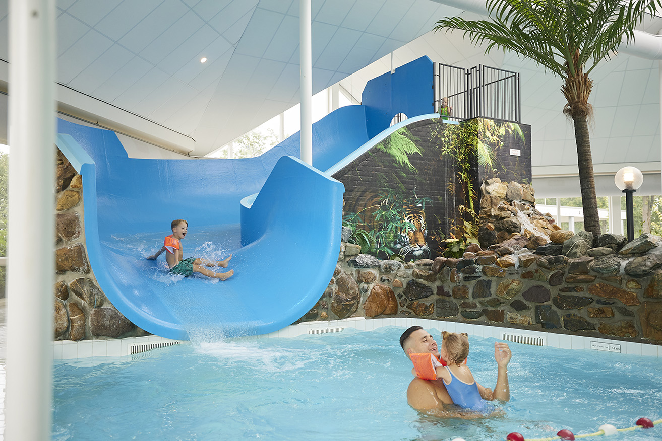 Een peuter glijdt van een brede blauwe glijbaan het kinderbad in van het subtropische zwembad op Recreatiepark De Leistert, waar een vader hem opwacht.
