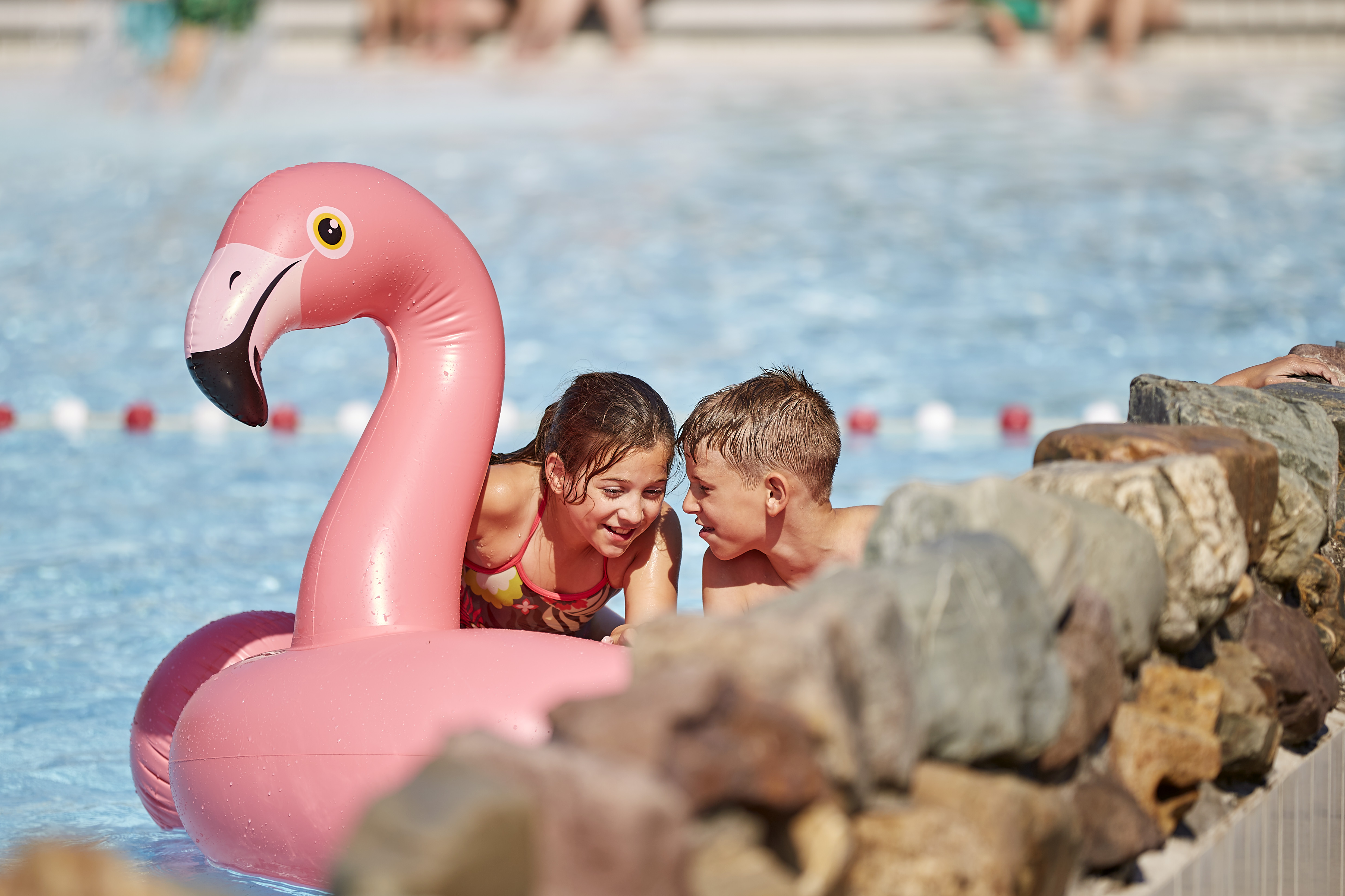 Twee kinderen fluisteren geheimpjes terwijl ze in het zwembad dobberen met een grote, roze, opblaasbare flamingo.