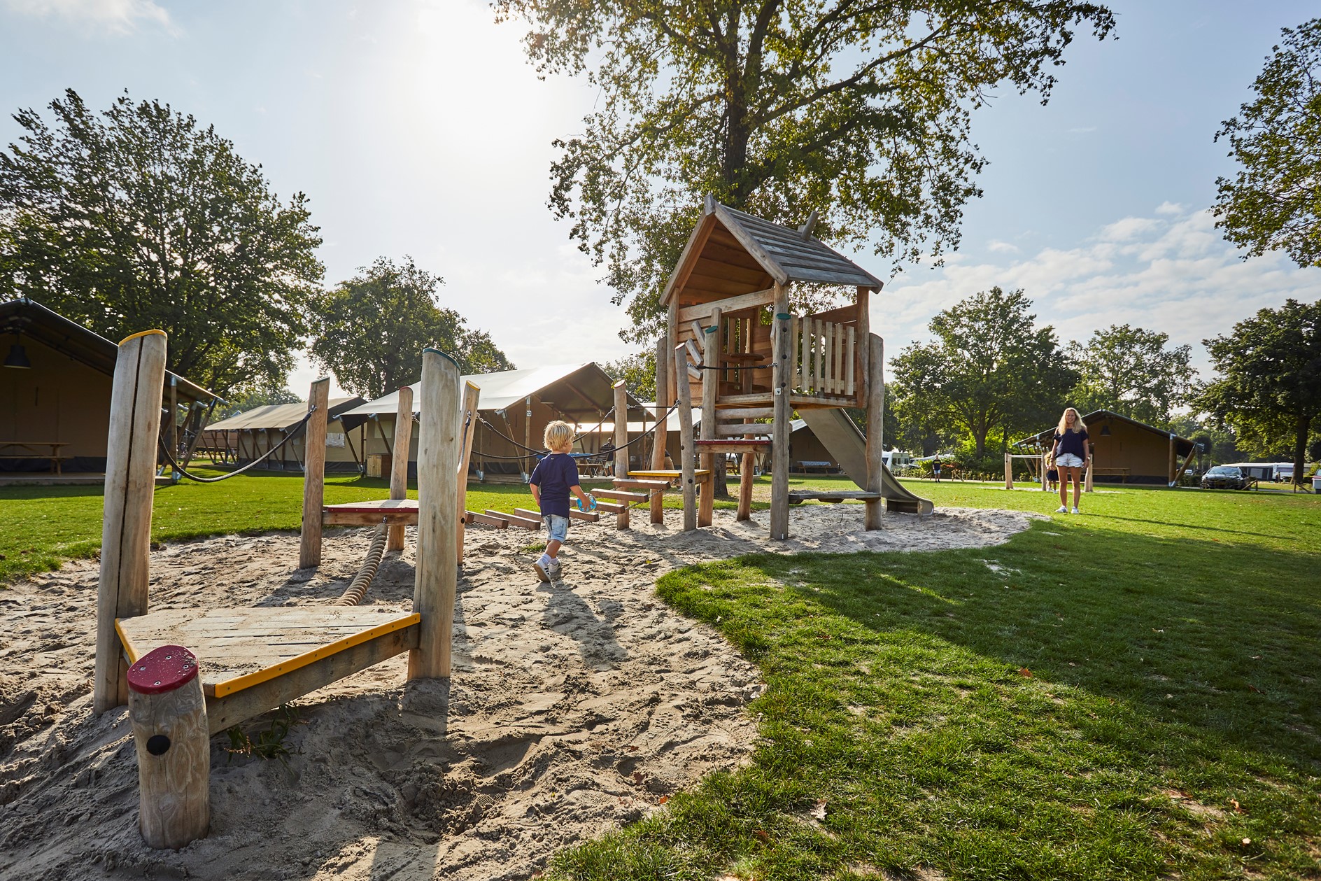 Een houten speeltoestel met glijbaan in een zandspeeltuin op Recreatiepark De Leistert, omgeven door een groot grasveld en bomen.