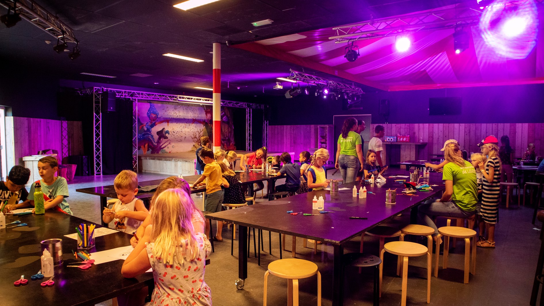 Een groep kinderen zit aan lange tafels te knutselen tijdens een activiteit van het animatieteam in de met paars licht verlichte theaterzaal van De Leistert.