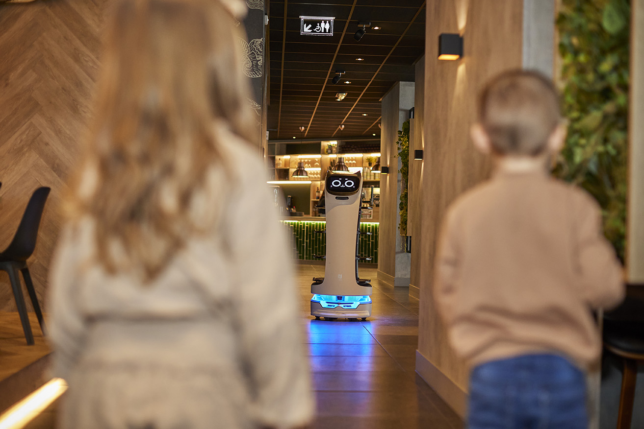 Twee kinderen kijken gefascineerd naar een serveerrobot die met blauwe lichten door de gang van een restaurant op De Leistert rijdt.