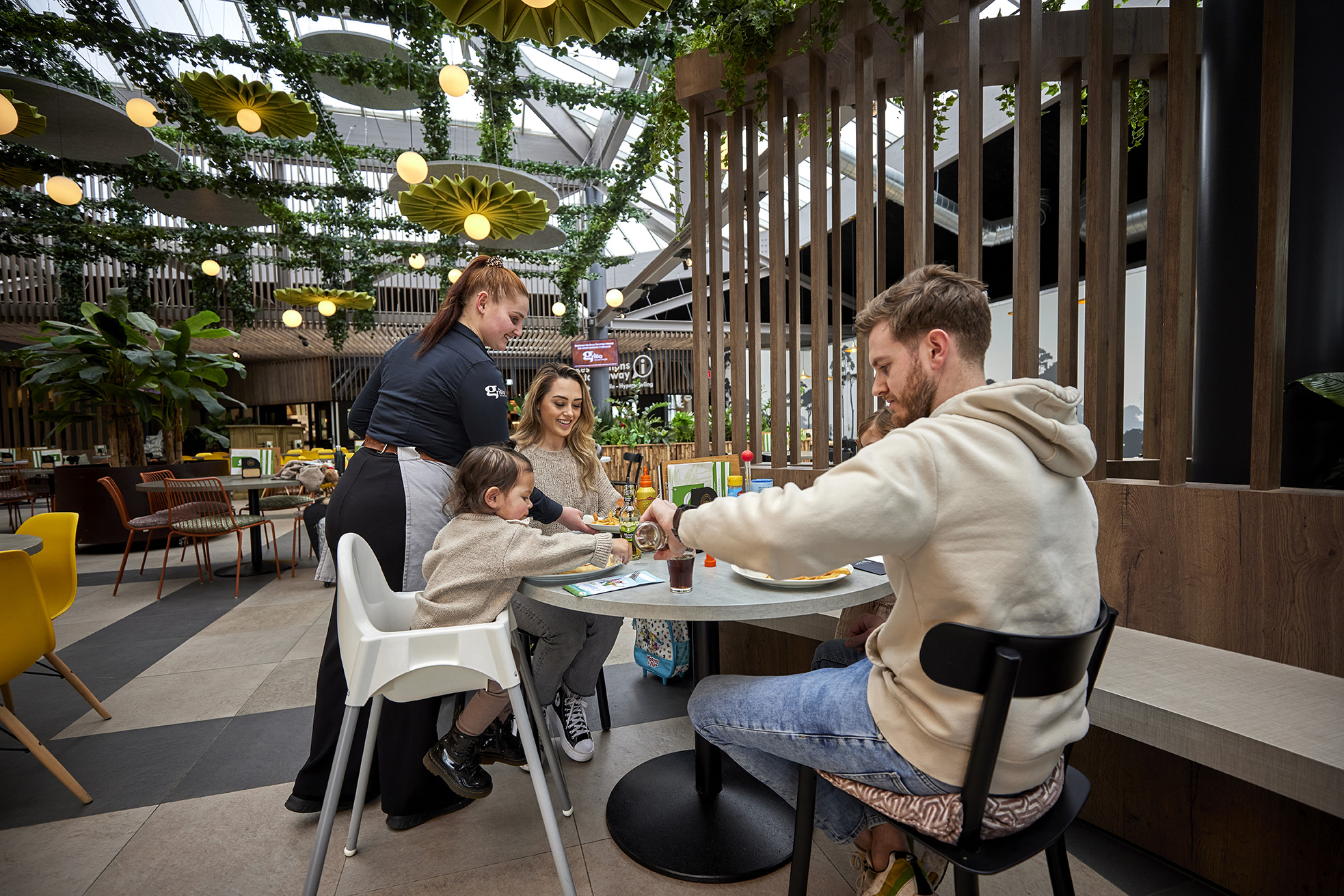 Een gezin met een kind in een kinderstoel zit te eten op het gezellige, overdekte terras van de Plaza op Recreatiepark De Leistert.