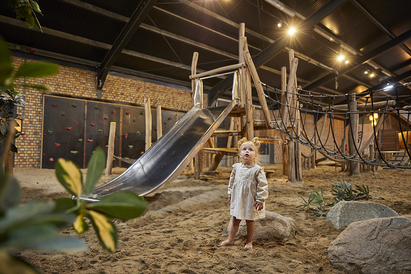 Een peuter staat in het zand van de indoor speeltuin La Selva op Recreatiepark De Leistert, naast een metalen glijbaan.