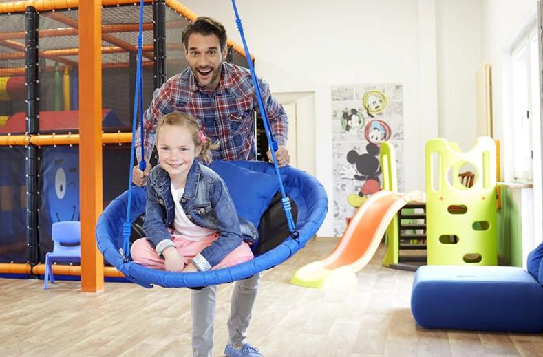 Een vader geeft zijn lachende dochter een duw op de grote schommel in de indoor speelruimte van Göbels Seehotel Diemelsee.