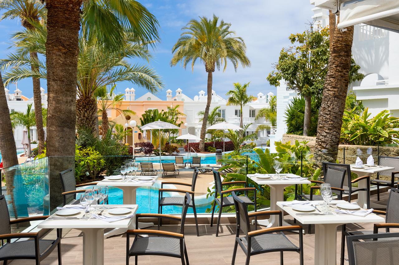 Gasten genieten van hun maaltijd aan tafels op het buitenterras van het restaurant, onder een pergola en met uitzicht op de tropische tuin.