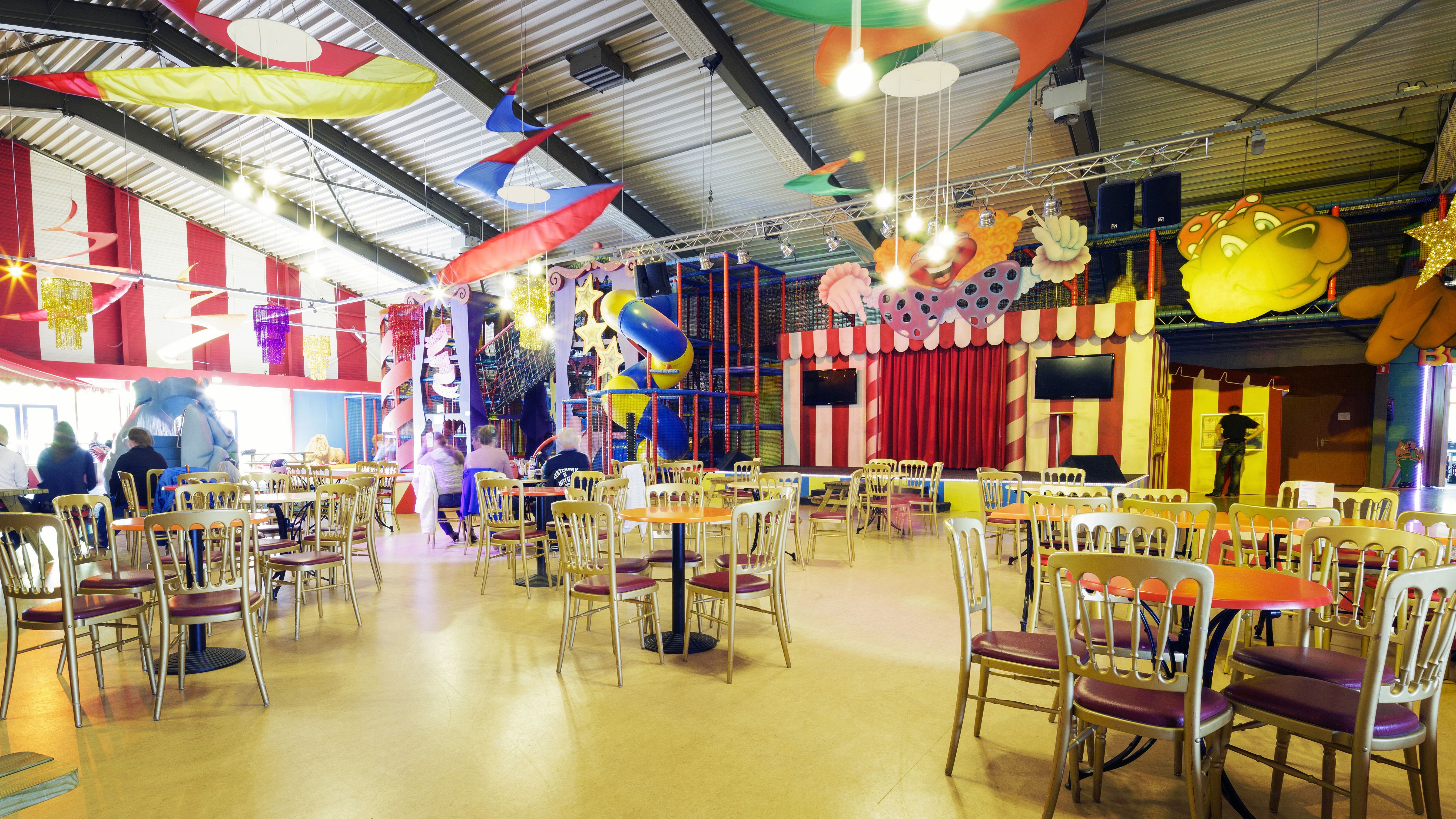 Het ruime en kleurrijke zitgedeelte voor ouders bij het indoorspeelparadijs van Landal Aelderholt, met uitzicht op de speeltoestellen.