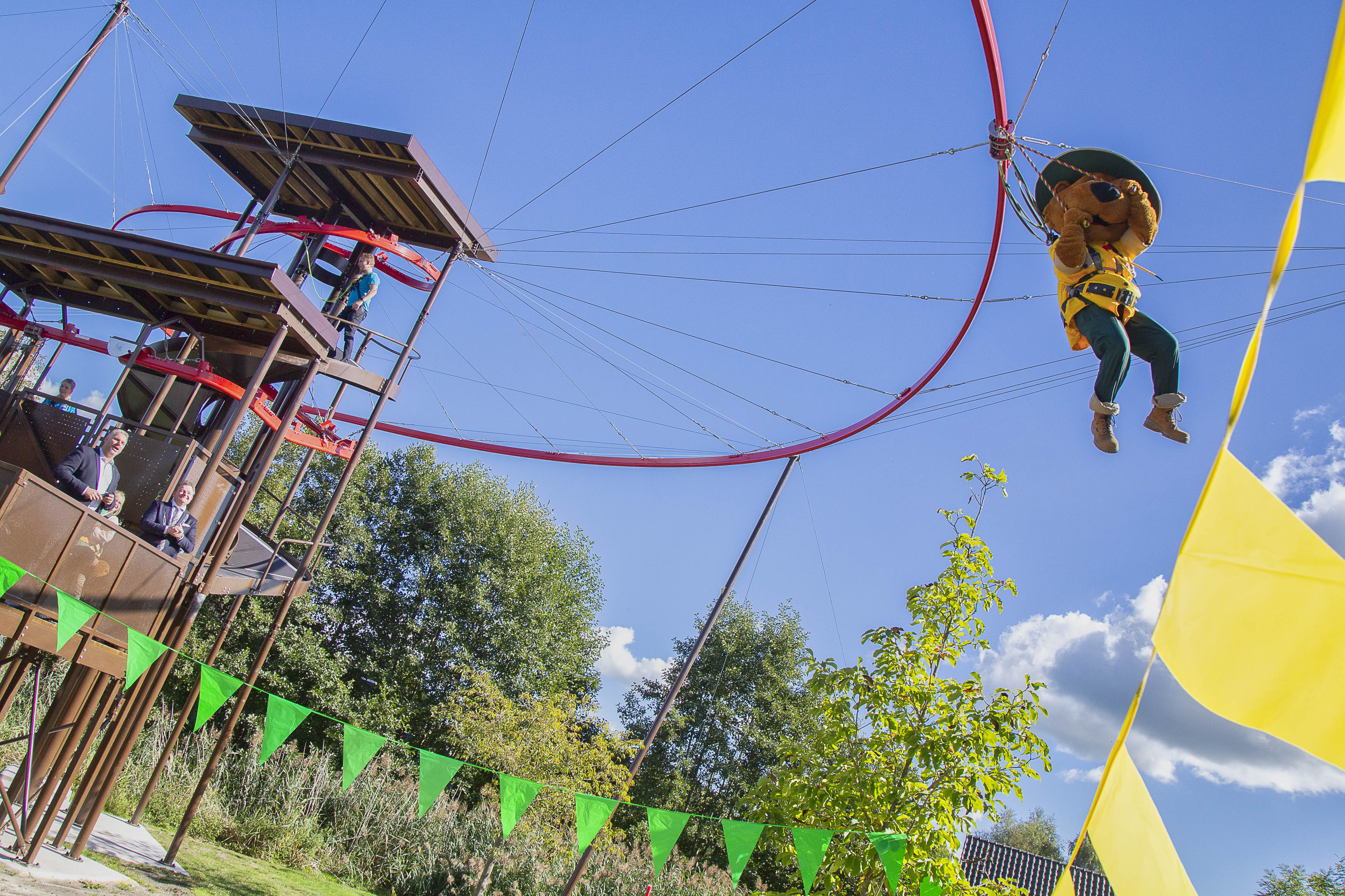 Een kind met een gele helm en veiligheidsharnas zoeft door de lucht aan de rode skyflyerl van het klimpark op Landal Aelderholt, met een blauwe lucht op de achtergrond.