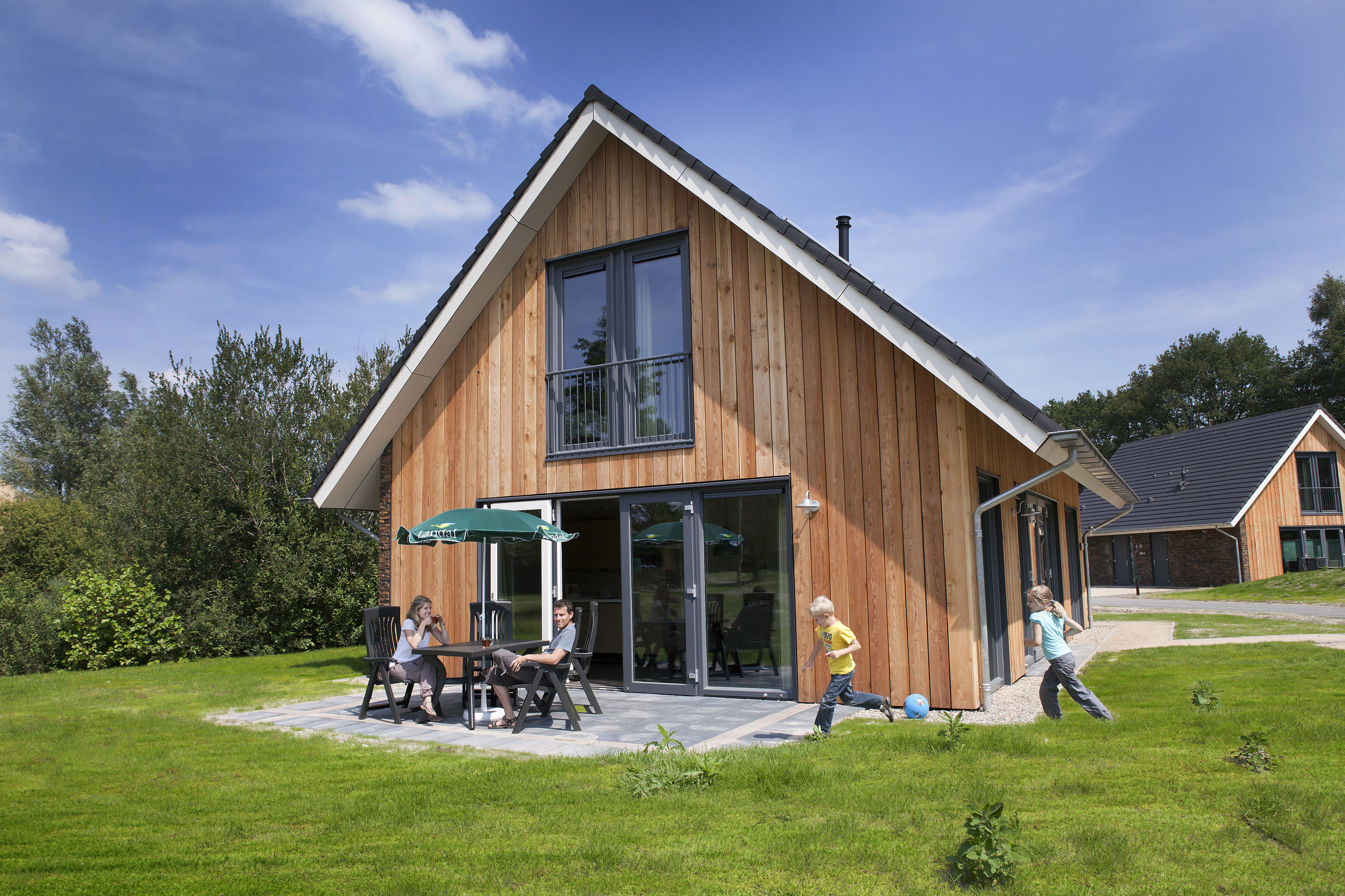 Een moderne, duurzame houten bungalow op Landal Aelderholt, waar ouders op het terras zitten en kinderen op het grasveld ervoor spelen op een zonnige dag.