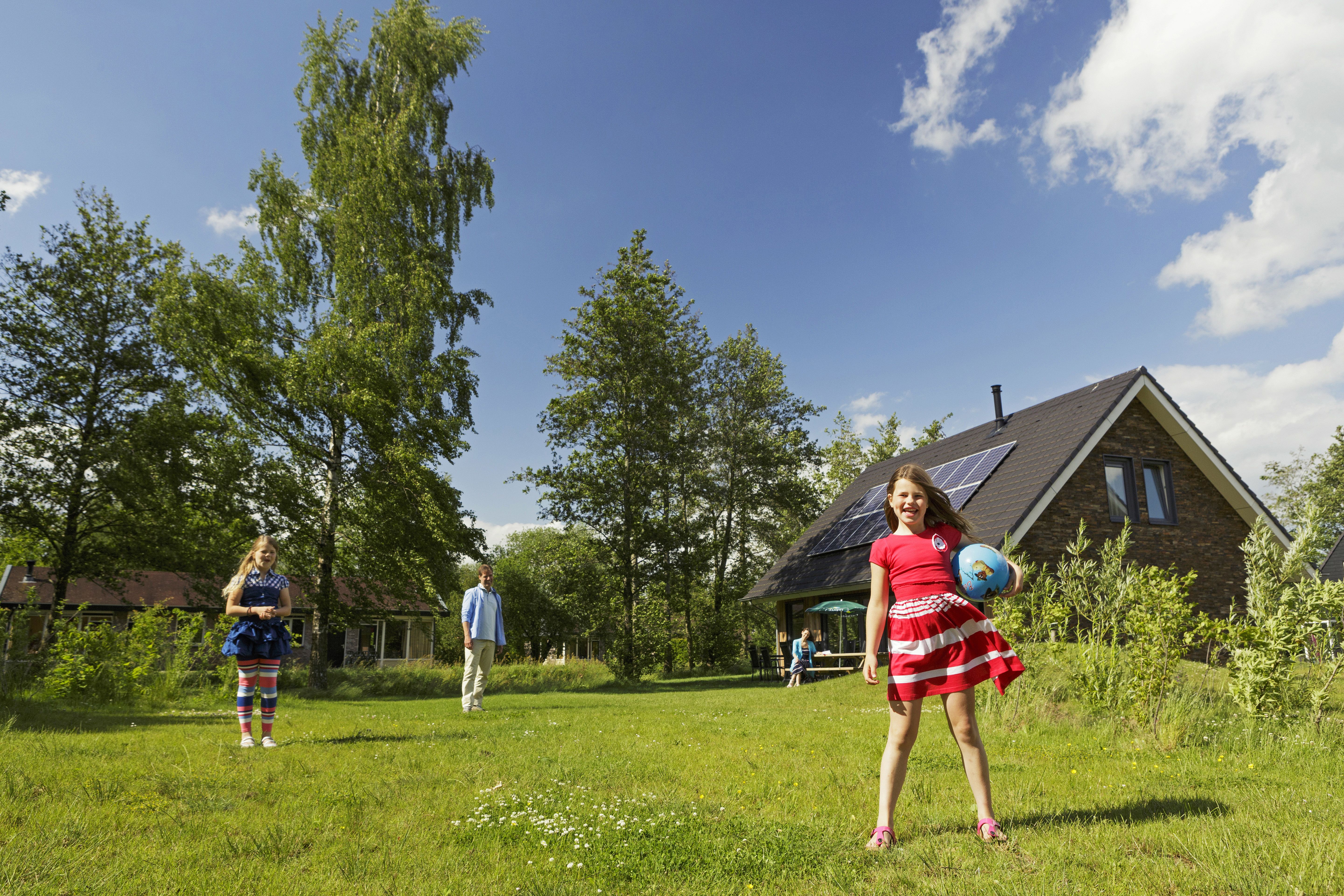 Drie kinderen spelen samen met een bal op een groot grasveld voor hun vakantiebungalow op een zonnige dag bij Landal Aelderholt.