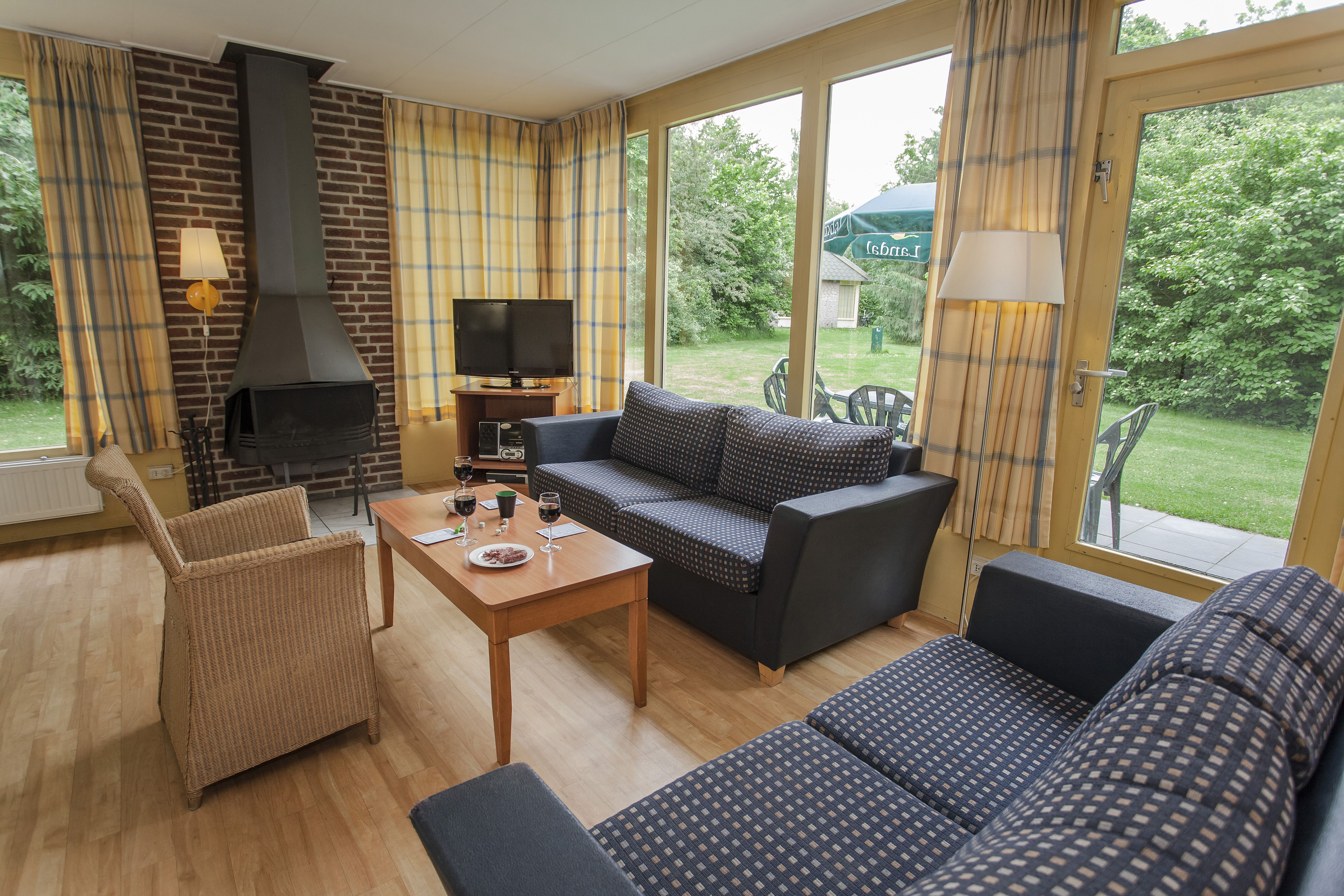 Een knus ingerichte woonkamer met een comfortabele zithoek, open haard en grote ramen met uitzicht op het groene buitengedeelte van de bungalow op Landal Aelderholt.