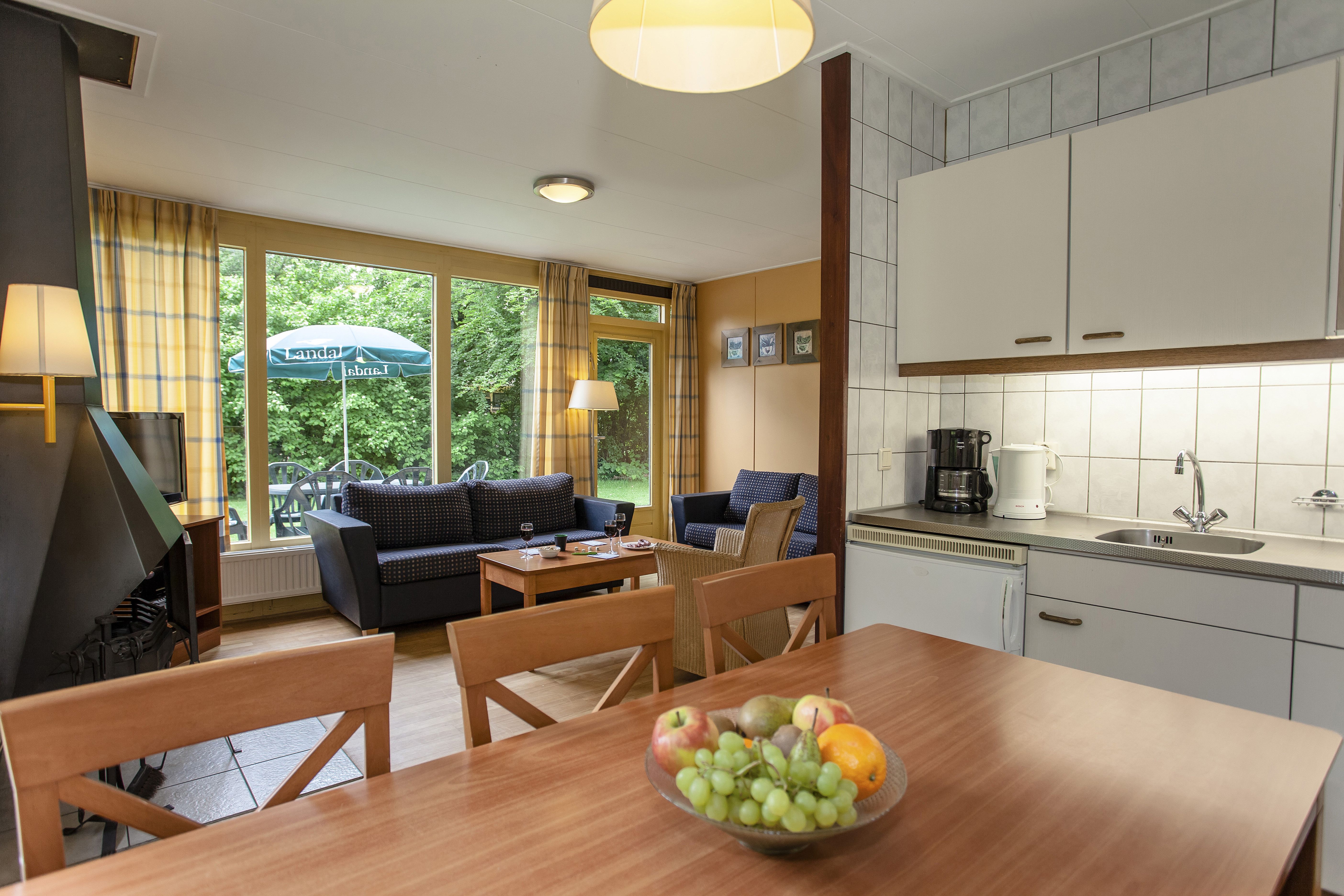 De open keuken en eethoek in een bungalow op Landal Aelderholt, met een houten eettafel en uitzicht op de zithoek en de groene tuin.