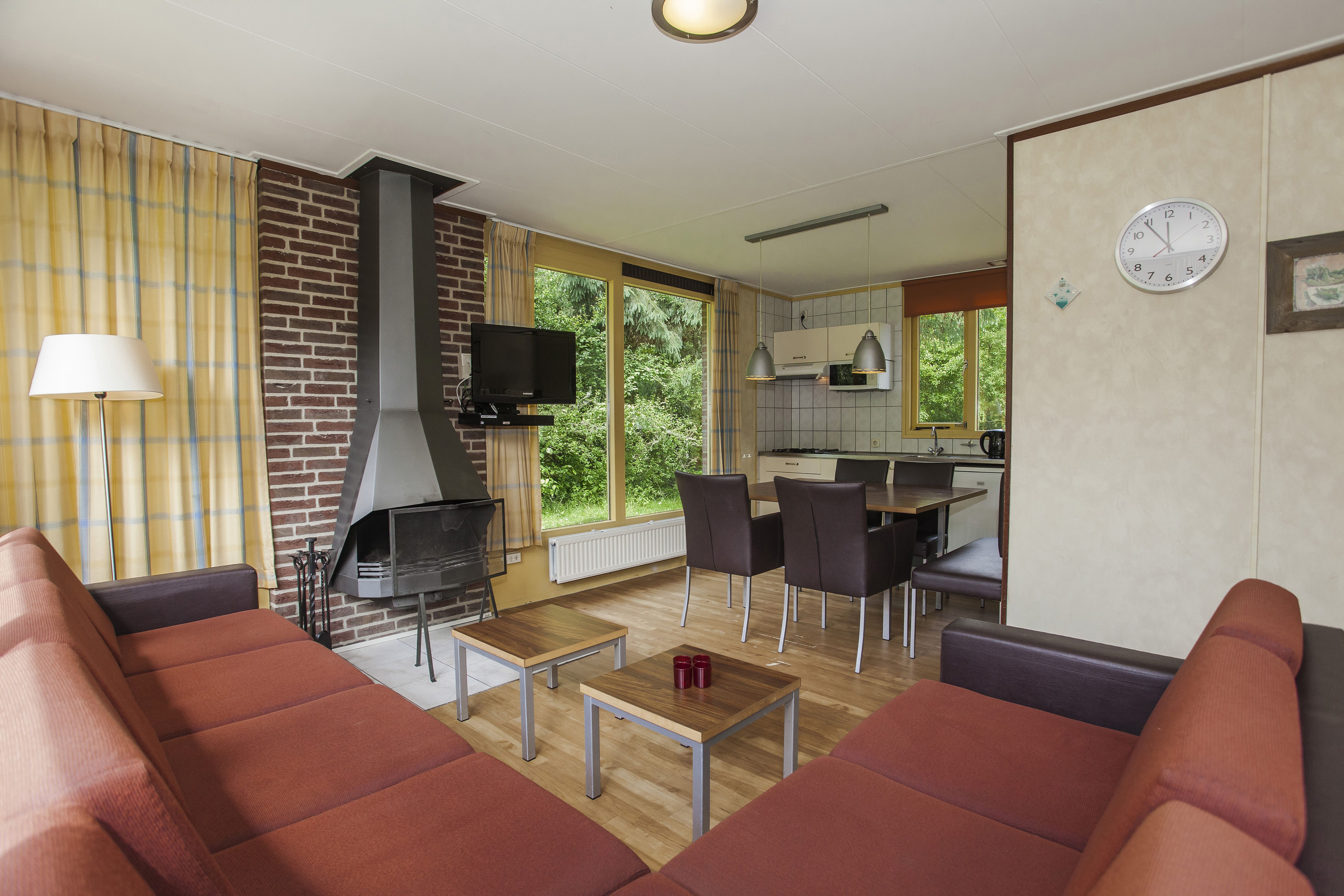 Een grote, open woonkamer van een groepsaccommodatie op Landal Sluftervallei, waar een familie ontspant op de banken en aan de lange eettafel.