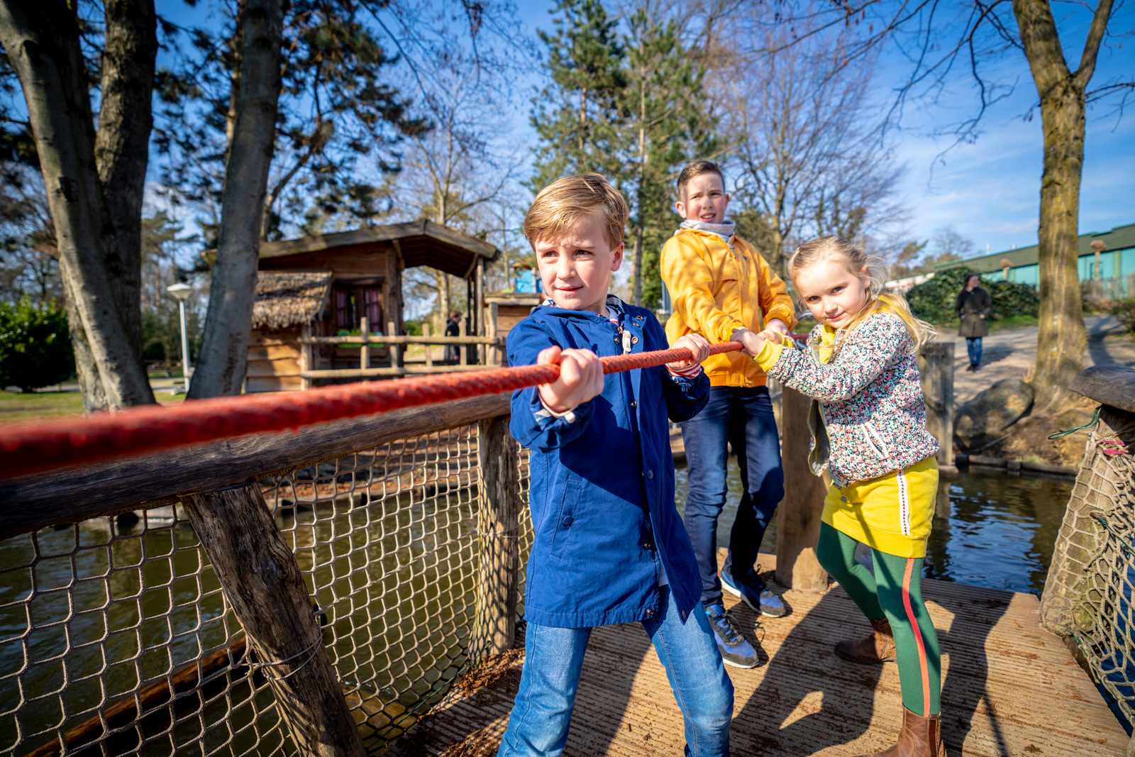 Drie kinderen werken samen en trekken aan een touw om een houten vlot over het water te bewegen in de avontuurlijke buitenspeeltuin van Landal De Lommerbergen.