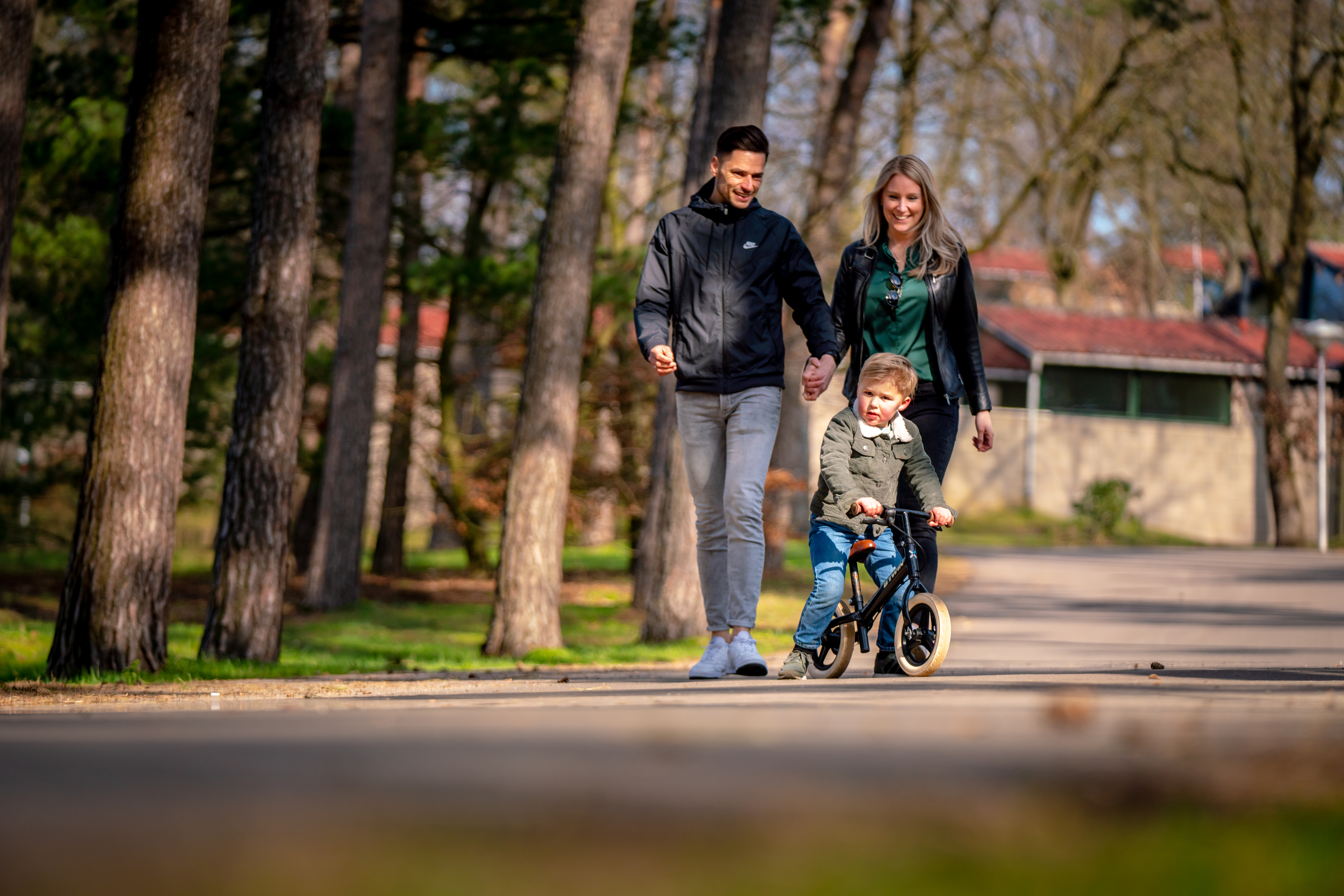Jong gezin wandelt hand in hand over een bospad op het autoluwe vakantiepark Landal De Lommerbergen, terwijl hun zoontje op een loopfiets rijdt.