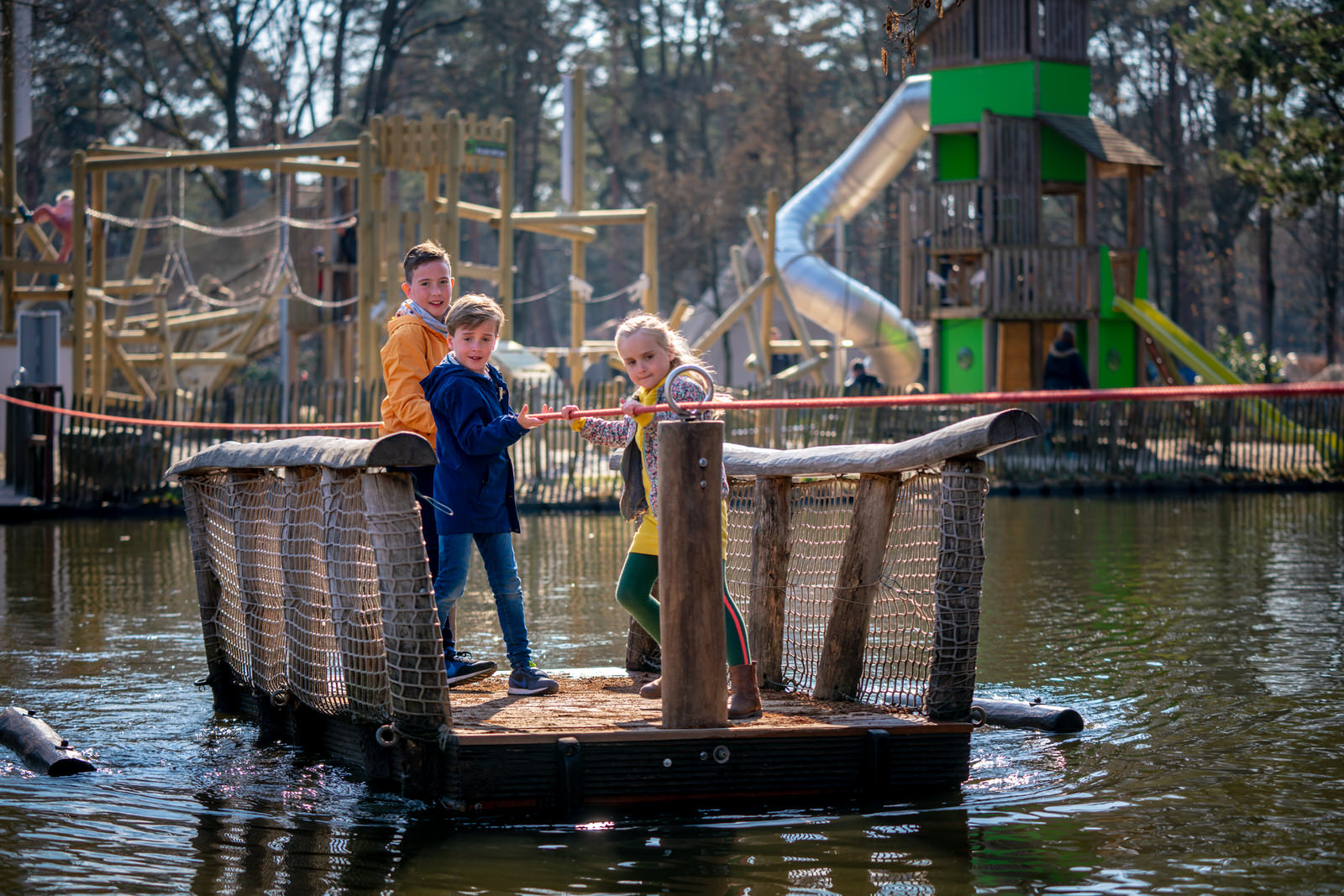 Kinderen op een houten trekvlot in de speelvijver met op de achtergrond de grote avontuurlijke buitenspeeltuin van Landal De Lommerbergen in Limburg.