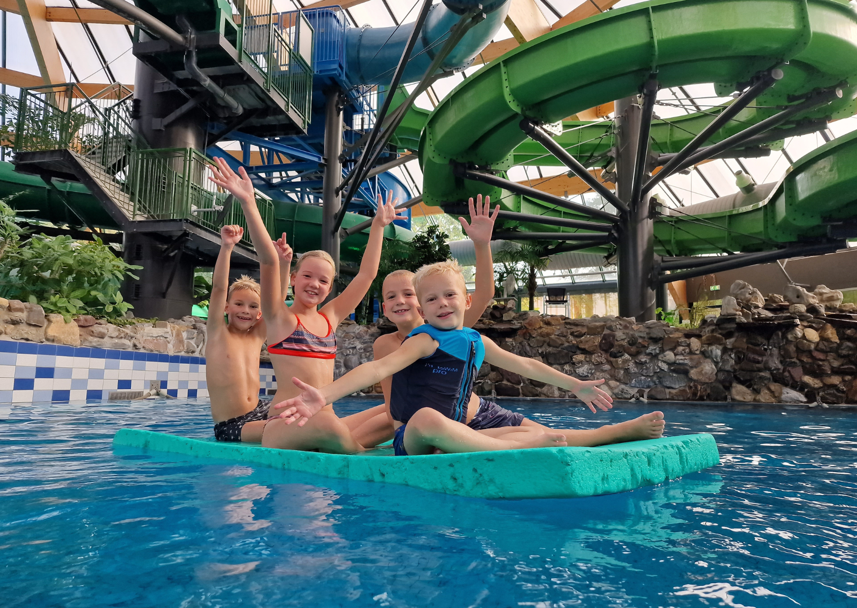 Vier vrolijk zwaaiende kinderen hebben plezier op een grote, drijvende mat in het zwembad van het subtropisch zwemparadijs op het kindvriendelijke vakantiepark Landal De Lommerbergen.