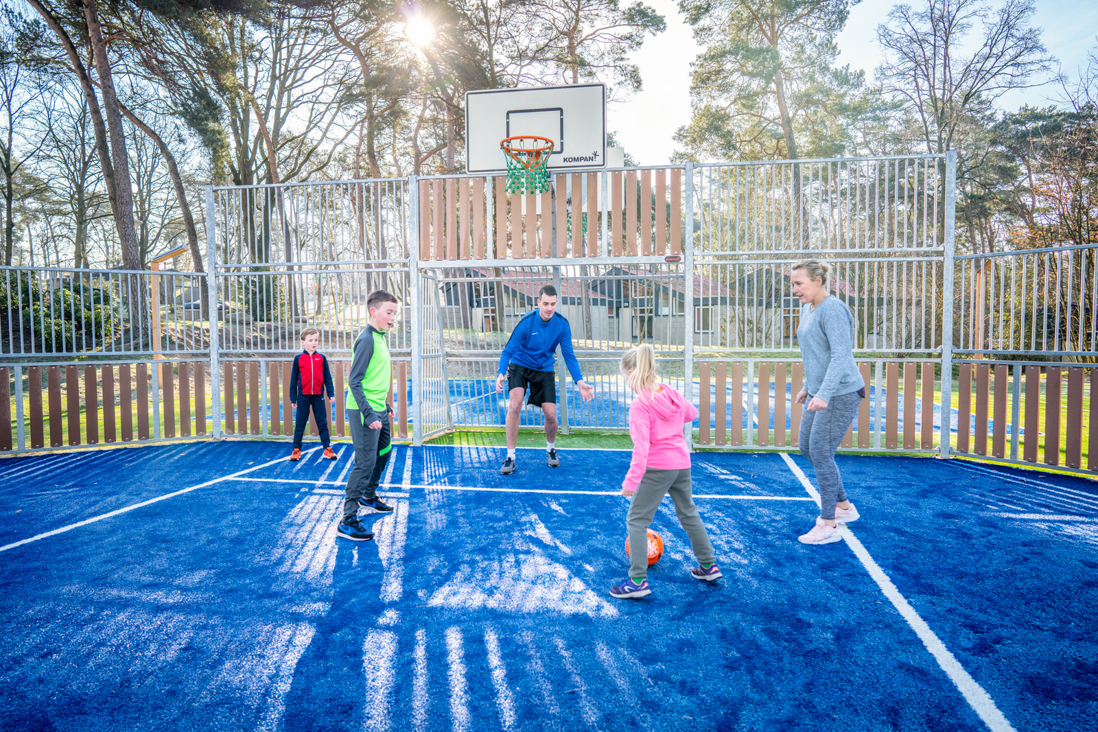 Kinderen en volwassenen spelen een potje voetbal en basketbal op het blauwe, multifunctionele sportveld op vakantiepark Landal De Lommerbergen.