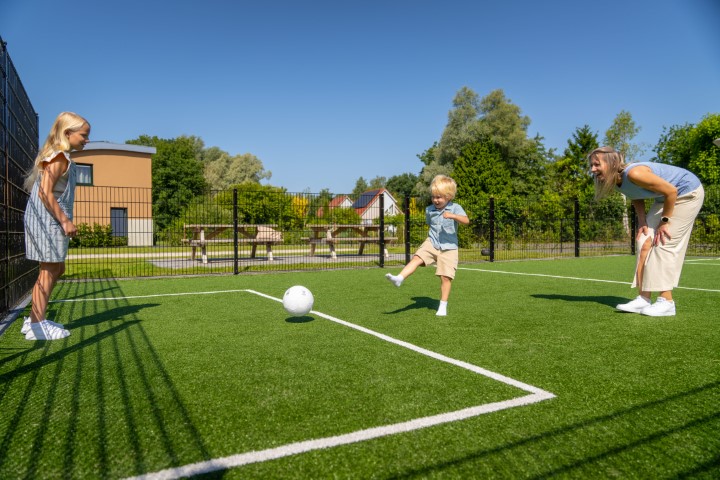 Een gezin, bestaande uit een moeder en twee kinderen, speelt enthousiast voetbal op het multifunctionele sportveld van Landal De Wedderplassen.