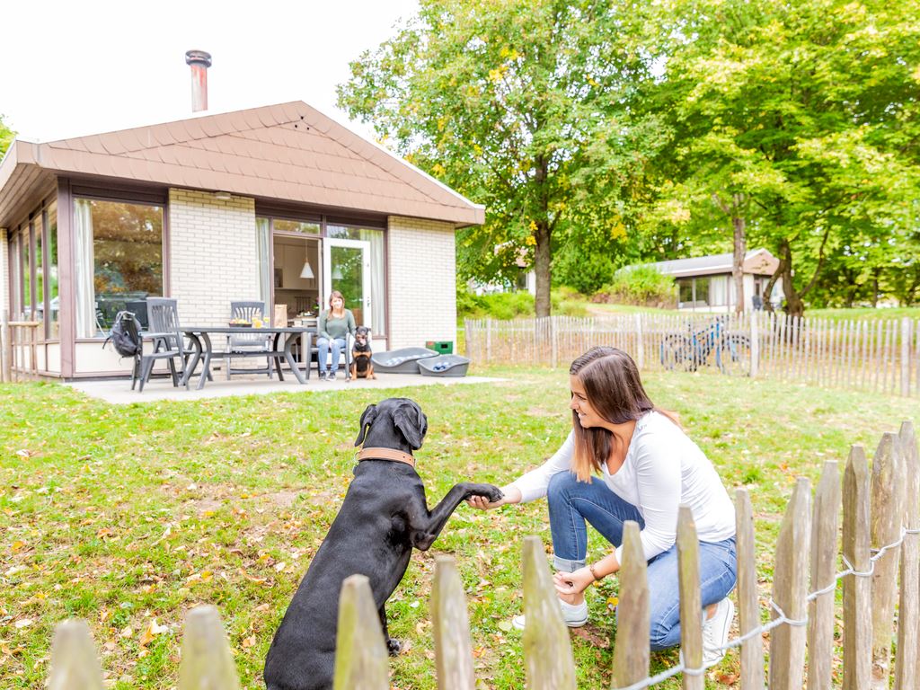 Een vrouw geeft een pootje aan haar zwarte hond in de omheinde tuin van een speciale hondenbungalow op Landal Hochwald.