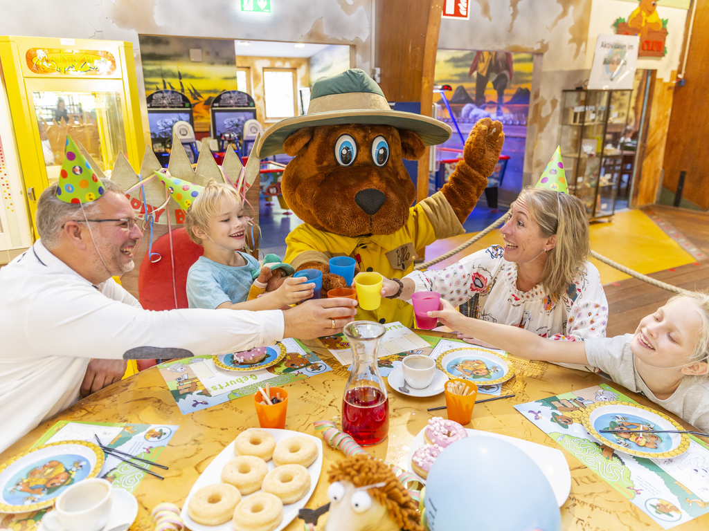 Een familie met kinderen viert een verjaardagsfeestje met mascotte Bollo en proost met feestmutsen op bij Landal Hochwald.
