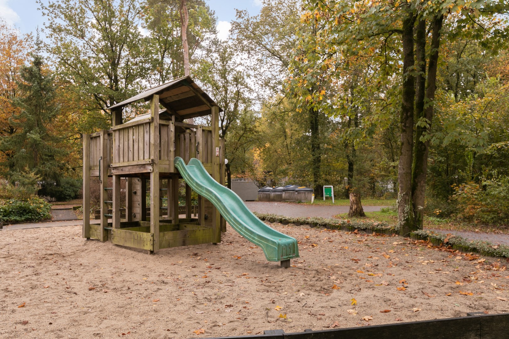 Een houten speeltoestel met glijbaan in een zandbak, gesitueerd in de bosrijke omgeving van Parc de Berkenhorst.