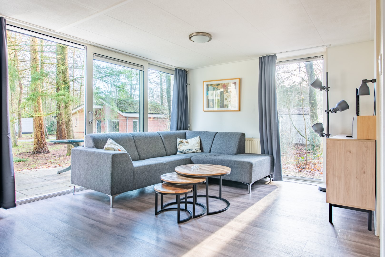 Lichte, moderne woonkamer met een grijze hoekbank en grote ramen die uitkijken op het bos bij Parc de Berkenhorst.