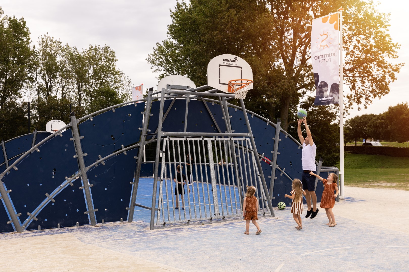 Kinderen en een volwassene spelen basketbal en voetbal in de multifunctionele sportkooi in het zand op Vakantiepark Port Greve.Kinderen en een volwassene spelen basketbal en voetbal in de multifunctionele sportkooi in het zand op Vakantiepark Port Greve.