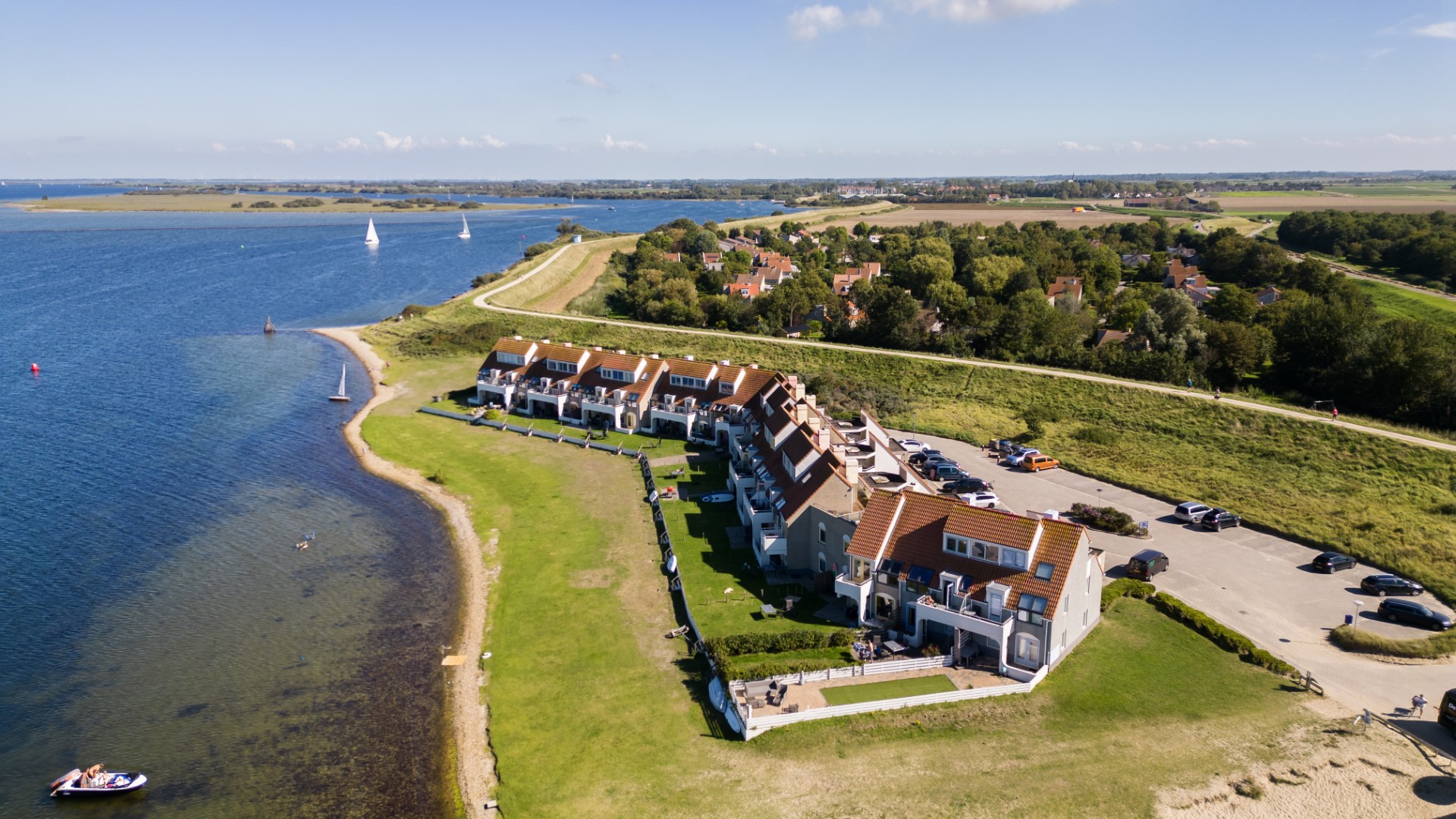 Een prachtige luchtfoto van Vakantiepark Port Greve aan het water in Zeeland, met een rij vakantiehuizen langs de kustlijn en zeilboten op het water.