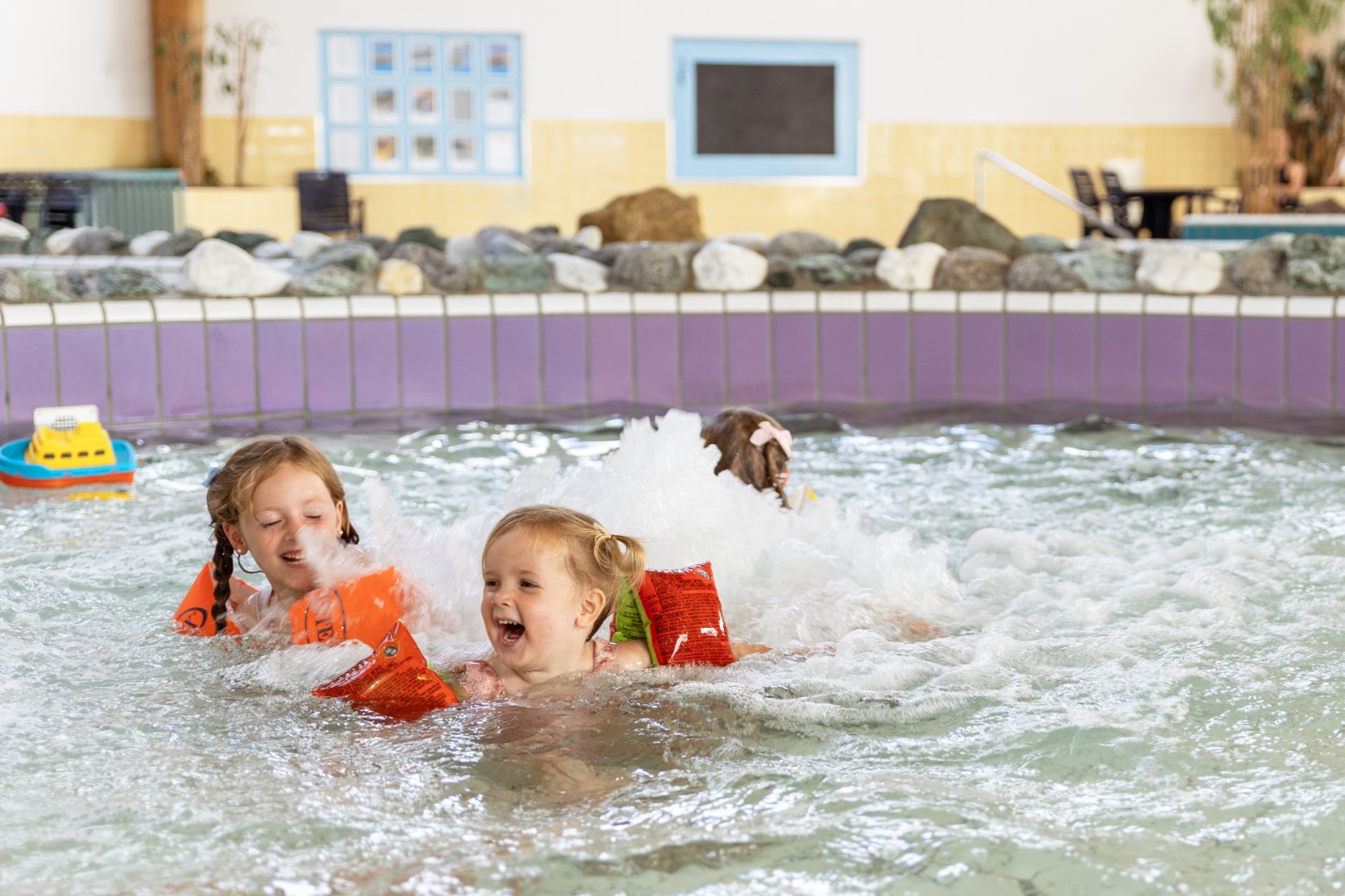 Twee lachende meisjes met zwembandjes spelen en spetteren in het ondiepe, bruisende kinderbad van het zwembad op Vakantiepark Port Greve.