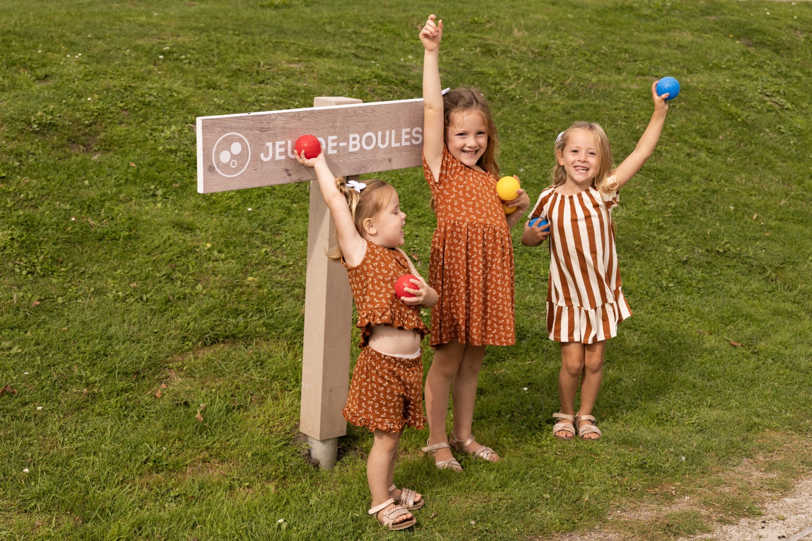Drie vrolijke zusjes staan met jeu-de-boulesballen in hun handen klaar voor een potje op de jeu-de-boulesbaan van Port Greve.