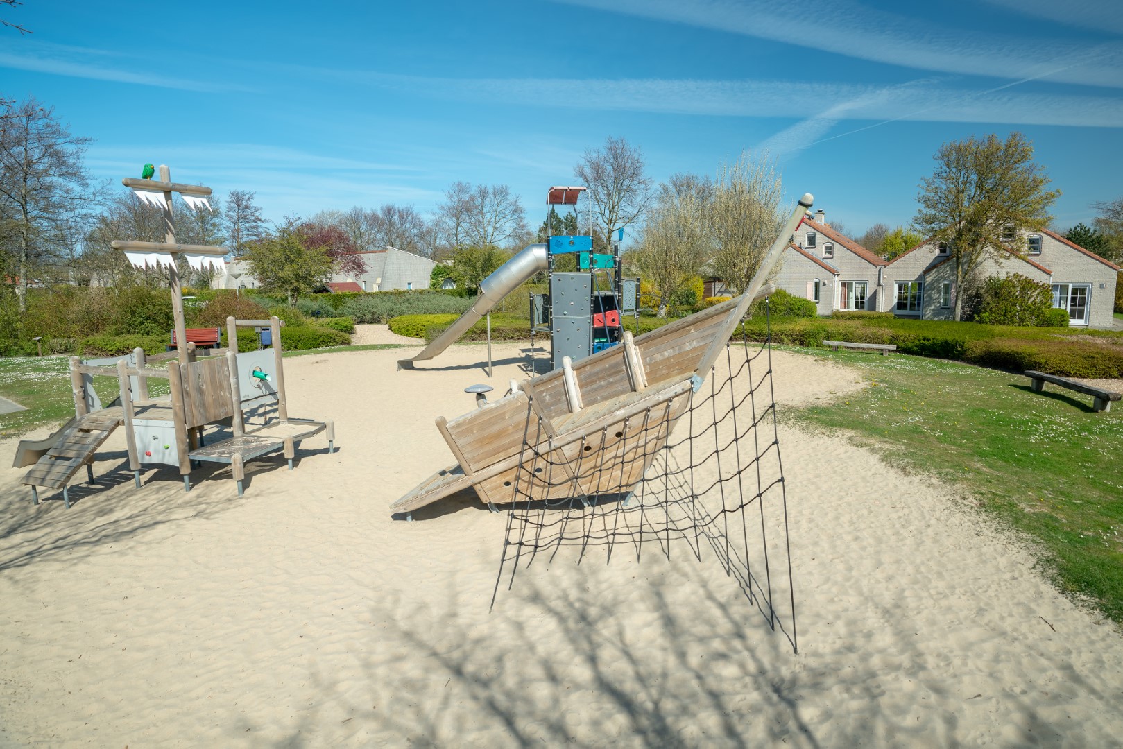 Een grote buitenspeeltuin in het zand op Vakantiepark Port Greve, met een houten piratenschip om op te klimmen en een glijbaan.