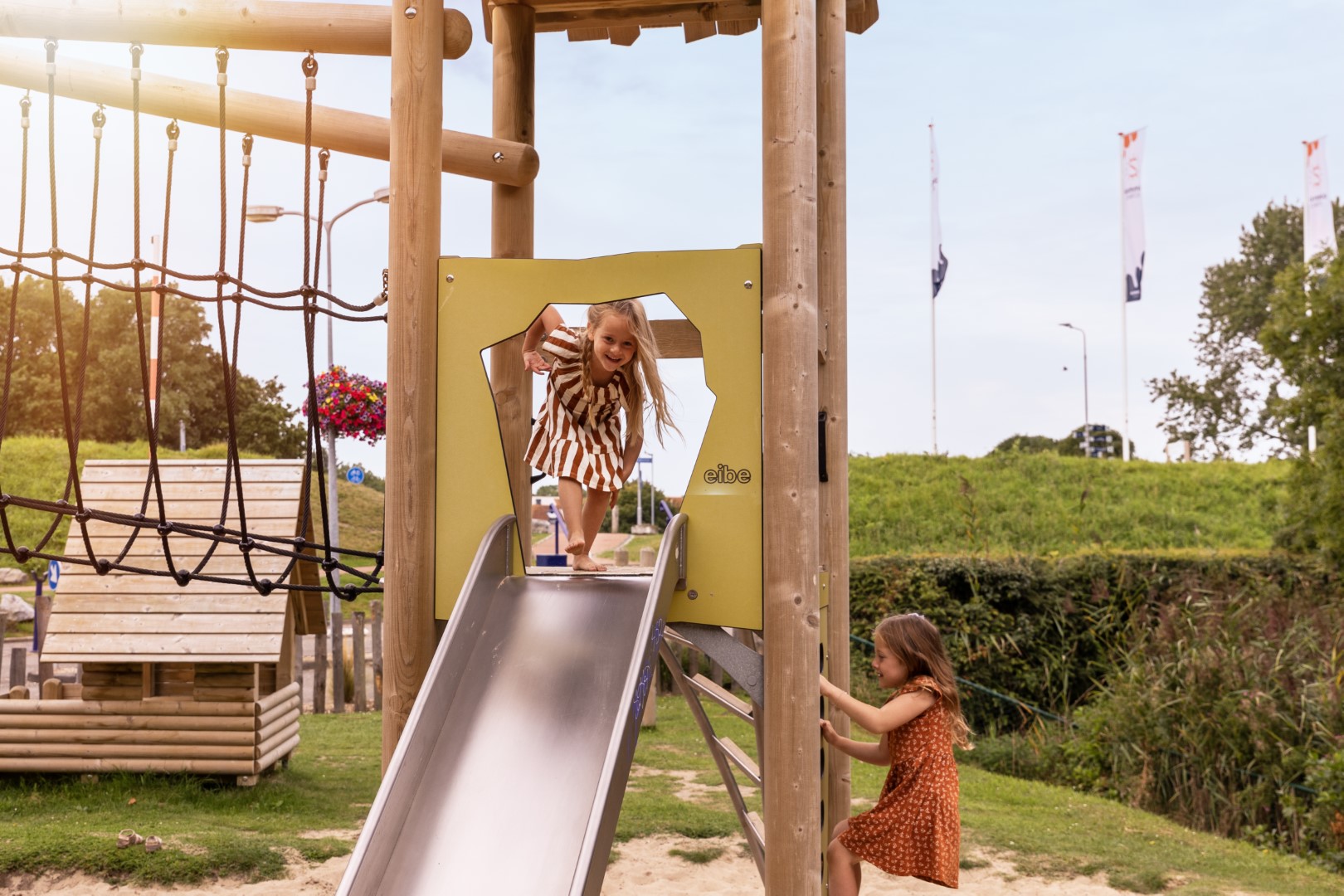 Twee meisjes spelen in de buitenspeeltuin van Vakantiepark Port Greve, een meisje glijdt van de glijbaan terwijl de ander toekijkt.