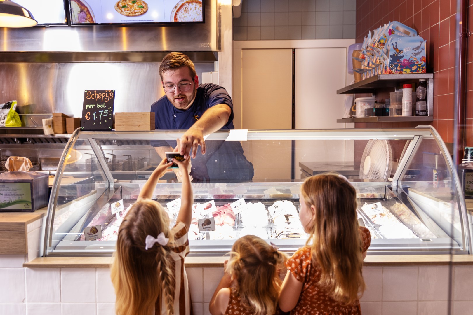 Drie meisjes staan bij de ijsvitrine van de snackbar op Vakantiepark Port Greve en krijgen een ijsje van een medewerker.