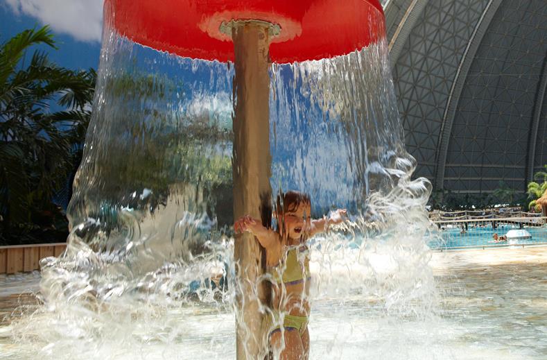 Een klein kind staat lachend onder een grote waterpaddenstoel die als een waterval van water naar beneden stort in het kinderbad van Tropical Islands.
