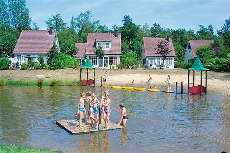 Kinderen spelen op een houten vlot in de zwemvijver van Vakantiepark Hellendoorn in Overijssel, met vakantiehuisjes op de achtergrond.