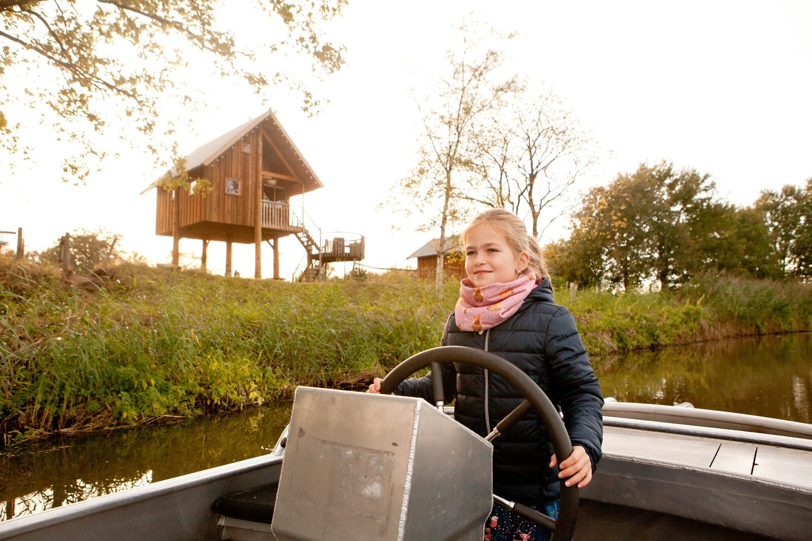 Een meisje staat aan het roer van een fluisterbootje en vaart over de rivier de Regge bij Vakantiepark Molke, met een boomhut op de achtergrond.